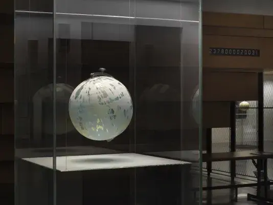 One painted globe suspended in a glass rectangular column and hangs over a flat surface that cuts the cylinder horizontally. Splatters of paint can be seen on top of the panel underneath the globe.