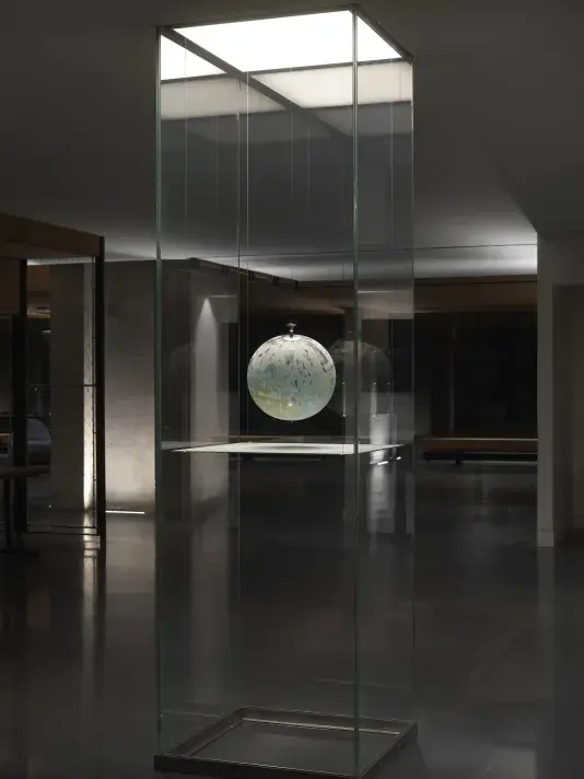 One painted globe suspended in a glass rectangular column and hangs over a flat surface that cuts the cylinder horizontally. Splatters of paint can be seen on top of the panel underneath the globe.