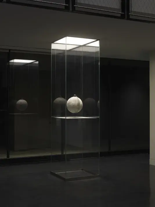 One painted globe suspended in a glass rectangular column and hangs over a flat surface that cuts the cylinder horizontally. Splatters of paint can be seen on top of the panel underneath the globe.