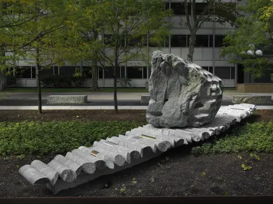 A large rock sculpture rests on a long strip of stone cylinders in a‬ ‭ greenspace. The rock has a series of holes removed from its core, with the‬ ‭ cylinders that were removed making up the strip it rests on. Trees, a pathway,‬ ‭ and a gray building reside in the background.‬