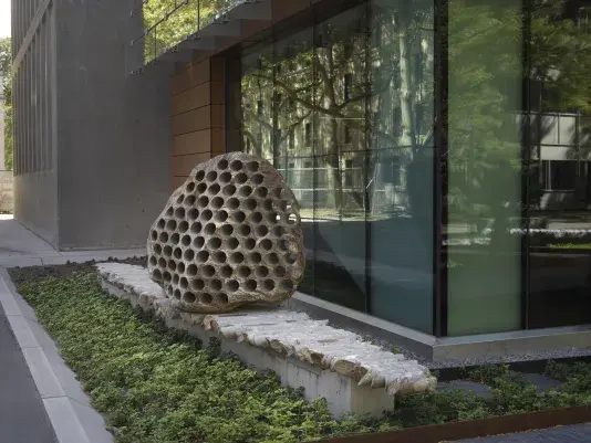 A large rock sculpture rests on a long strip of stone cylinders in a‬ ‭ greenspace next to the corner of a building. The rock has a series of holes‬ ‭ removed from its core, with the cylinders that were removed making up the strip it‬ ‭ rests on.‬