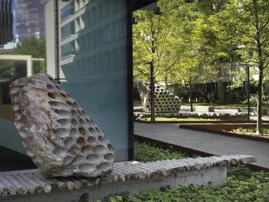 Two large rock sculptures rest on long strips of stone cylinders in a green space next to the corner of a building. each rock has a series of holes removed from its core, with the cylinders that were removed making up the strip it rests on. One sculpture is located in the lower left corner of the image, with the other in the background surrounded by trees.