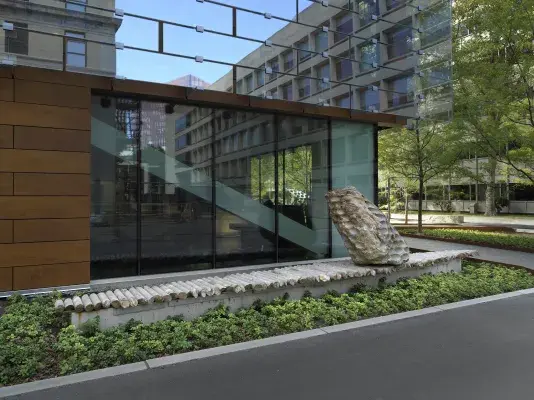 A large rock sculpture rests on a long strip of stone cylinders in a‬ ‭ greenspace next to the corner of a building. The rock has a series of holes‬ ‭ removed from its core, with the cylinders that were removed making up the strip it‬ ‭rests on.‬ ‭