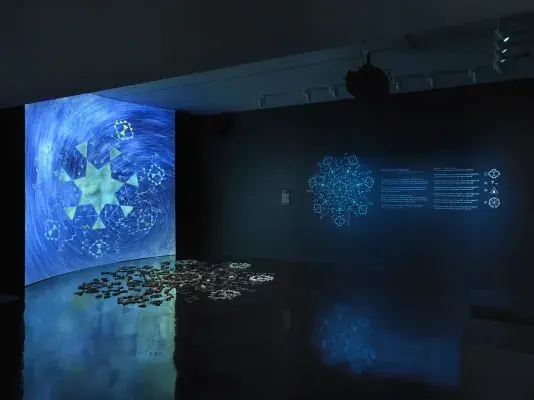 A dimly lit gallery space bathed in blue light. On the left, a large screen‬ ‭ displays a green six-pointed star at its center, surrounded by six inward-facing‬ ‭ triangles. Additional geometric shapes swirl into the blue background. In front of the‬ ‭ screen, a formation of stones rests on a reflective black floor. On the right wall,‬ ‭glowing blue geometric forms and wall text are visible.‬ 
