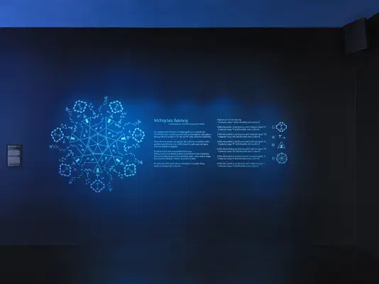 On a dark blue gallery wall, a glowing circle of geometric forms made up of‬ ‭ Lakhota characters is displayed. To its right, instructions for a score commissioned‬ ‭ by the American Composers Orchestra are present. Further to the right, four‬ ‭ Lakhota characters are shown, representing the elements that compose the‬ ‭geometric form on the left.‬ 