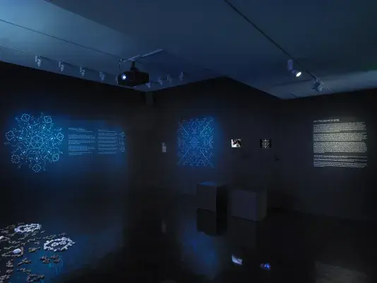 A dimly lit gallery space glows blue. Symbols illuminate the back walls, while‬ ‭ exhibition wall text and iPads displaying video footage are positioned on the far right.‬ ‭ In the lower left corner, a formation of stones peeks into the frame.‬ 