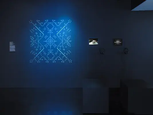 In a dark gallery space, a blue-glowing geometric form is shown on the wall.‬ ‭To its right, two small iPads showcase video footage—one featuring a drum and the‬ ‭other displaying a musical score.‬ 