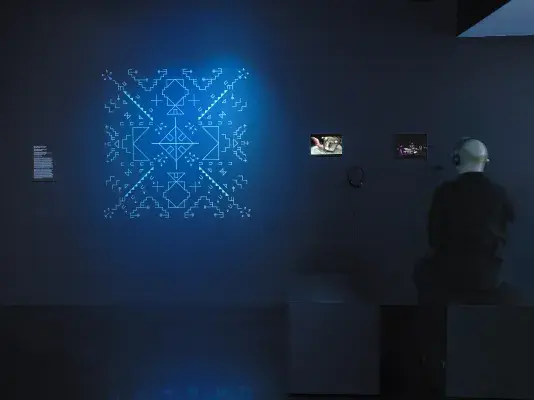 In a dark gallery space, a blue-glowing geometric form is displayed on the‬ ‭ wall. To its right, two small iPads show video footage—one featuring a drum and the‬ ‭ other displaying a musical score. On the far right, a man wearing headphones sits,‬ ‭watching one of the iPads.‬ ‭