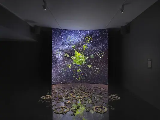 A dark gallery space features a large screen displaying a six-sided star at its‬ ‭ center. Within the star, video footage of green stones is projected. Surrounding the‬ ‭ star, six inward-facing triangles form a pattern, while geometric shapes swirl in the‬ ‭ background alongside footage of stars and the night sky. In front of the screen, a‬ ‭formation of stones rests on the reflective black floor.‬ ‭