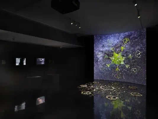 A dark gallery space bathed in tinted blue light. On the right, a large screen‬ ‭ displays a six-sided star at its center, with video footage of green stones projected‬ ‭ within it. Surrounding the star, six inward-facing triangles form a pattern, while‬ ‭ geometric shapes swirl in the background alongside footage of stars and the night‬ ‭ sky. In front of the screen, a formation of stones rests on the reflective black floor.‬ ‭ On the left wall, two videos of performers dancing are displayed.‬