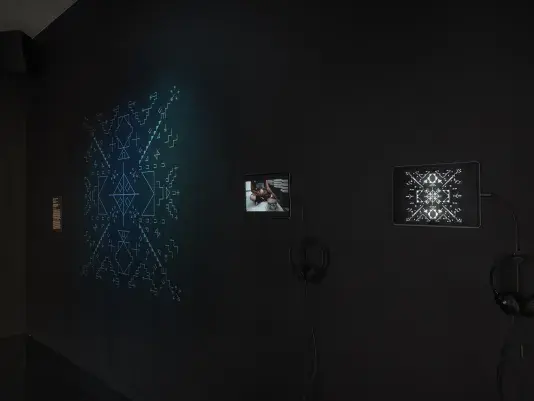 A dimly lit gallery space with two iPads are on display. The far right iPad‬ ‭ shows white geometric forms on a black background, and to its left the other iPad‬ ‭ shows a person drumming. On the far left, a dimly lit form made up of geometric‬ ‭symbols glows blue.‬