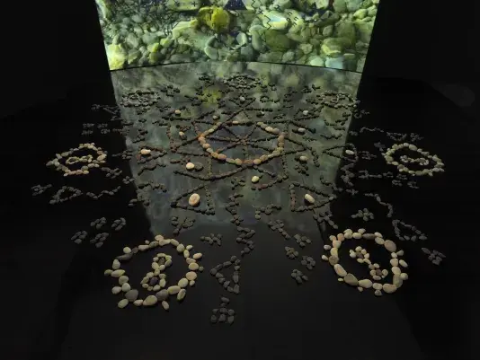 In a dark gallery space, a formation of stones rests on a black mirrored floor.‬ ‭ The stones are placed in different formations, circles, zig-zags, triangles, infinity‬ ‭ symbols, which all meet at a central circular point. Behind the stones is a large‬ ‭ screen showing video footage of rocks under water. The video footage reflects off‬ ‭the mirrored floor.‬‭