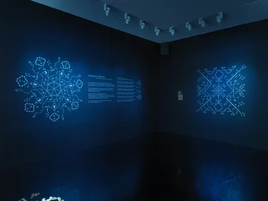 In the corner of a dark blue gallery wall, two circles of geometric symbols‬ ‭ made up of Lakhota characters glow on either side of the corner. Between the two‬ ‭ formations of geometric symbols are instructions for a score commissioned by the‬ American Composers Orchestra.‬