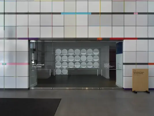 Entryway with glass doors surrounded by a geometric gray paneled wall with colorful accents. A black and white video projection of a series of images of the sun in a grid can be seen through the glass doors.