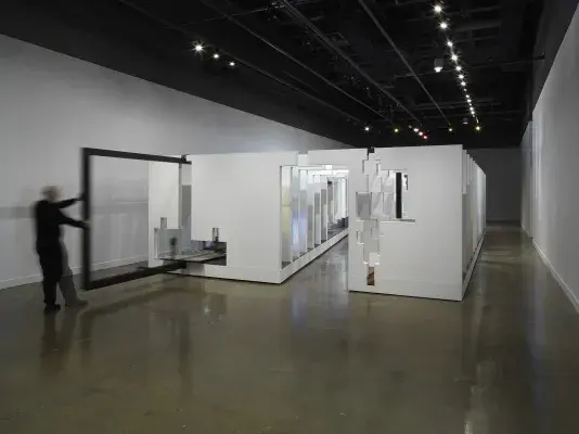 In the middle of an open space, there is a large structure with a series of fragmented white walls divides a living space with wooden floors, furniture, and house plants throughout. Each section of wall is connected by black beams. A man in a all black pulls one of the black beams on the left side of the frame.