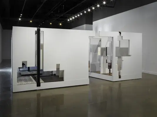 In the middle of an open space, there is a large structure with a series of fragmented white walls divides a living space with wooden floors, furniture, and house plants throughout. Each section of wall is connected by black beams.