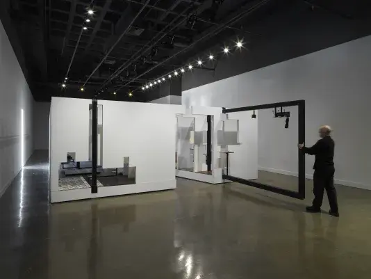 In the middle of an open space, there is a large structure with a series of fragmented white walls divides a living space with wooden floors, furniture, and house plants throughout. Each section of wall is connected by black beams. A man in a all black pulls one of the black beams on the right side of the frame.