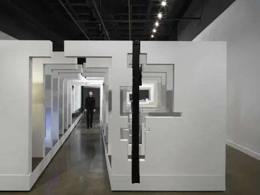 A series of fragmented white walls divides a living space with wooden floors, furniture, and house plants throughout. Each section of wall is connected by black beams. A woman in a all black walks through an opening in the middle of the structure.