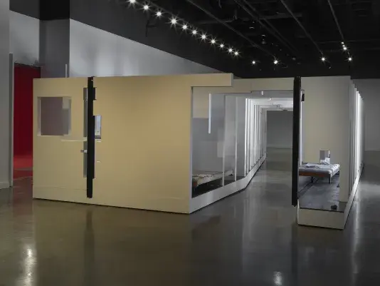 In the middle of an open space, there is a large structure with a series of fragmented white walls divides a living space with wooden floors, furniture, and house plants throughout. Each section of wall is connected by black beams.