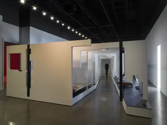 A series of fragmented white walls divides a living space with wooden floors, furniture, and house plants throughout. Each section of wall is connected by black beams. A woman in a all black walks through an opening in the middle of the structure.