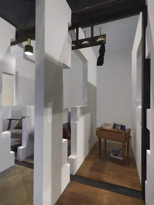 A series of fragmented white walls divides a living space with wooden floors, furniture, and house plants throughout. A wooden nightstand with a stereo and a stack of books is at the center of the frame. Each section of wall is connected by black beams.