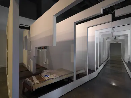 A walkway surrounded on both sides by a series of white walls that segments what appears to be a living space. A charging smartphone is visible on top of a bed in the bottom lefthand corner.