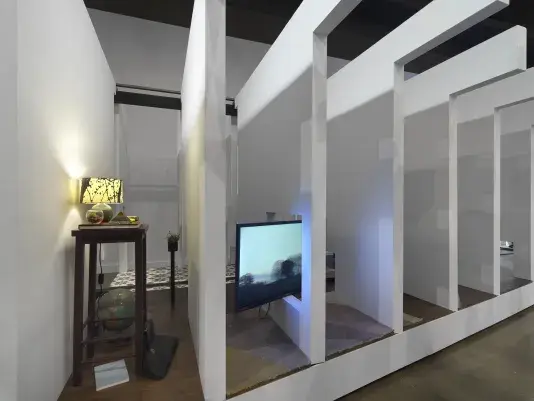 A series of fragmented white walls divides a living space with wooden floors, furniture, and house plants throughout. Each section of wall is connected by black beams. There is a wooden nightstand with a lamp on top and a globe on the ground beside at the left side of the frame. A flatscreen TV is divided by a white wall on the right side of the frame.