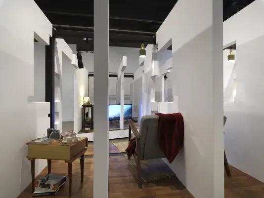 A series of fragmented white walls divides a living space with wooden floors, furniture, and house plants throughout. Each section of wall is connected by black beams. There is a wooden nightstand with a stack of books on top at the left side of the frame. The back of a couch is visible divided by a white wall on the right side of the frame.