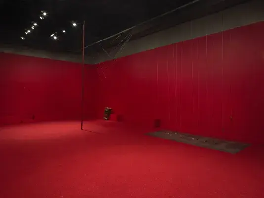 A copper poll stands vertical in the middle of a completely red painted walled room with red carpeting. To the right side there are a series of weights on strings above a metal plate. A wooden cabinet tilted on a angle is visible against the wall behind. Three metal spheres hang on strings suspended from the ceiling throughout the room.