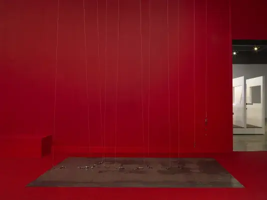 A series of weights suspended on strings above a metal plate in the middle of a completely red painted walled room with red carpeting. On the right side there is a doorway to bright room.