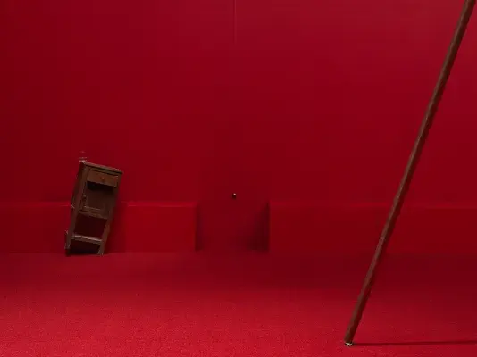 A wooden cabinet standing on a angle tilted to the right in a completely red painted room with red carpeting, next to a metal sphere hanging on a string suspended from the ceiling. Part of a copper poll is angled to the right in the right hand side of the frame.