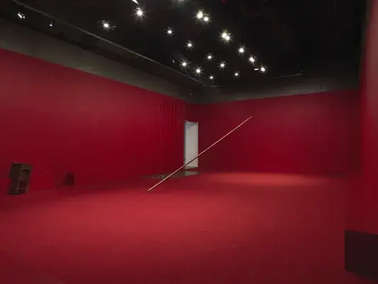 A copper poll suspended on an angle in the middle of a completely red painted walled room with red carpeting. Behind there are a series of weights on strings above a metal plate. A wooden cabinet tilted on a angle is visible against the wall to the left of the frame. Metal spheres hang on strings suspended from the ceiling throughout the room.