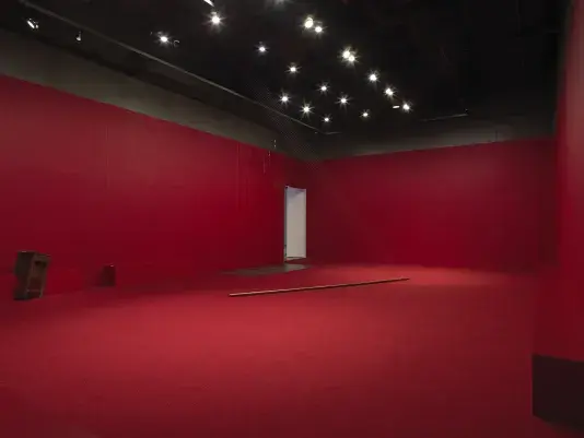 A copper poll suspended on an angle in the middle of a completely red painted walled room with red carpeting. Behind there are a series of weights on strings above a metal plate. A wooden cabinet tilted on a angle is visible against the wall to the left of the frame. Metal spheres hang on strings suspended from the ceiling throughout the room.