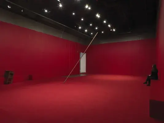 A copper poll suspended on an angle in the middle of a completely red painted walled room with red carpeting. Behind there are a series of weights on strings above a metal plate. A wooden cabinet tilted on a angle is visible against the wall to the left of the frame. A woman in all black sits on rectangular block at the edge of the room to the right side of the frame.
