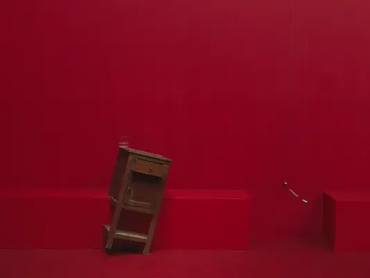 A wooden cabinet tilted on a angle to the right in a completely red painted room with red carpeting. A swinging metal sphere on a string suspended from the ceiling is captured in motion.
