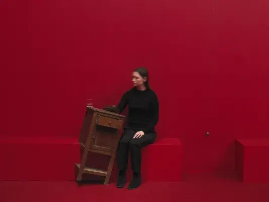 A wooden cabinet tilted on a angle to the right in a completely red painted room with red carpeting. A woman in a black sits to the right with her hand inside of the cabinet. A swinging metal sphere on a string suspended from the ceiling is captured in motion.