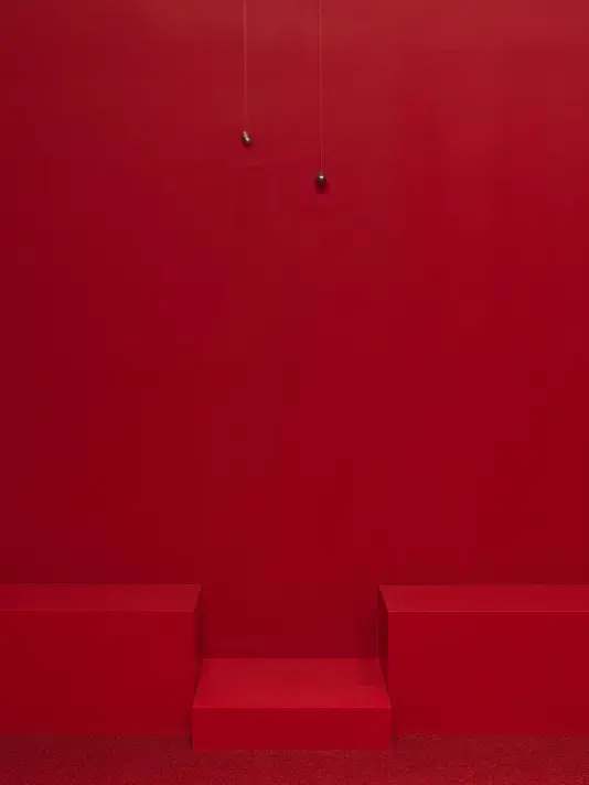 Two swinging metal spheres suspended from the ceiling on strings are captured in motion in a completely red painted walled room with red carpet.