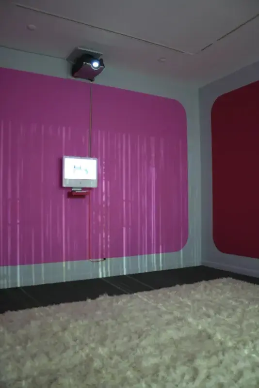 2007 Mac computer monitor affixed to a wall with a pink rectangle with a plush white rug on the ground. 