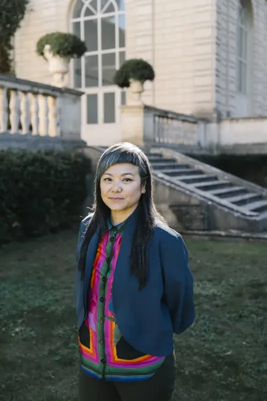 A portrait of Lan Tuazon, a woman with a light skin tone and long black and grey hair. She stands outside with her hands behind her back wearing a soft smile and a brightly colored button-up and blazer.