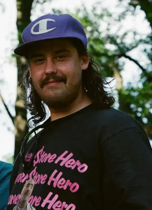Portrait of Adam Khalil, a man of the Ojibway tribe with medium-light skin tone and black shoulder-length hair softly smiling wearing a blue Champion baseball cap and black shirt.