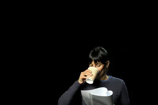 Portrait of Amy Balkin, a woman with dark hair and medium-light skin tone looking down at a packet of paper while drinking out of a paper coffee cup.