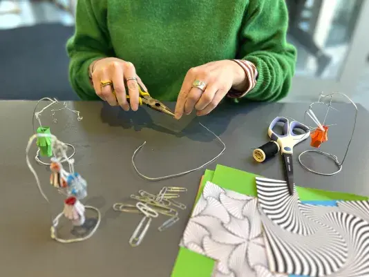Two hands bending wire with wire cutters. There is a small mobile with a green paper lantern to the left side of the hands, a group of paper clips and a stack of unfolded paper in front of the hands, and scissors with another wire mobile to the right of the hands.