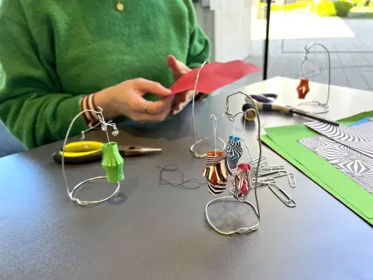 Two hands folding a red piece of paper. There is a small mobile with a green paper lantern to the left side of the hands, a group of paper clips and a stack of unfolded paper in front of the hands, and scissors with another wire mobile to the right of the hands.