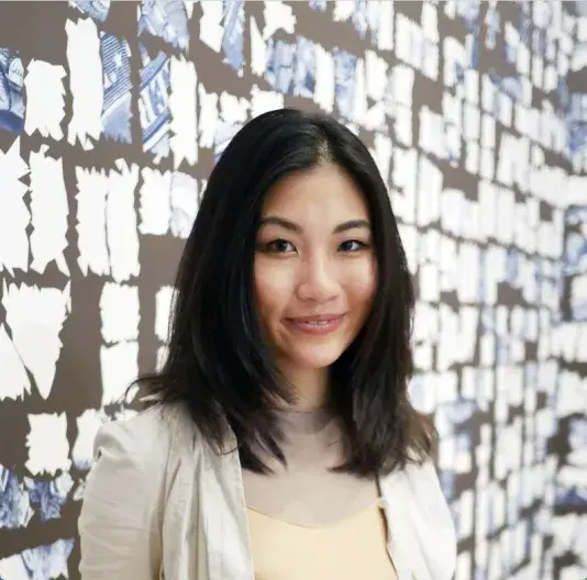 Portrait of Beatrice Glow, an American woman of Taiwanese heritage with medium-light skin tone and black shoulder-length hair softly smiles in front of a wallpapered wall from her art installation.