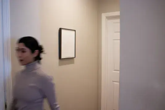 A work of art in a black frame hanging on a wall to the left of a white door. To the left of the artwork the blurry figure of a woman is walking out of the corridor.