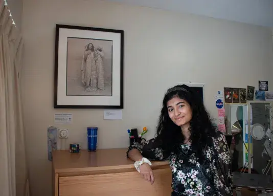 Woman leaning on a light wooden dresser wearing a floral printed top. To the left of the woman there is a artwork in a black frame hanging on the wall.
