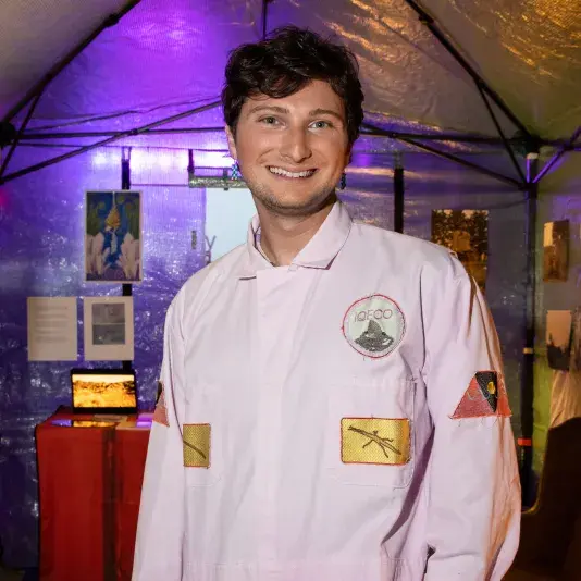 A portrait of Lee Pivnik, a man with a light skin tone and short brunette hair. He stands in a tent with artwork pinned behind, in the foreground he smiles broadly wearing a pale pink jumpsuit with fraying iron-on patches.