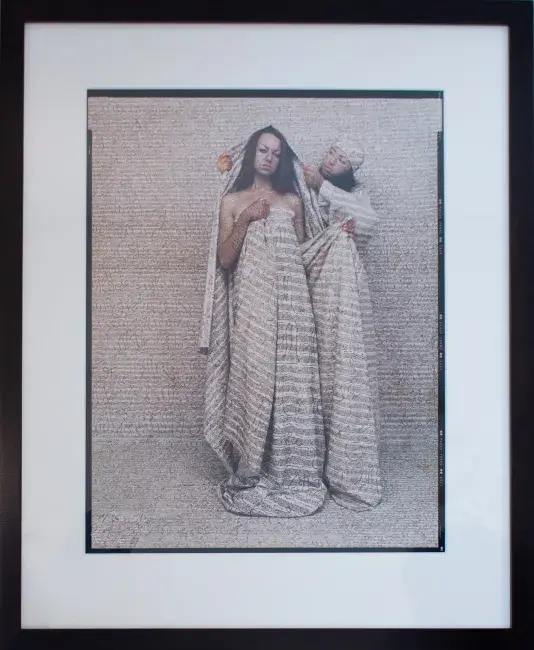 Two women standing in front of a beige background covered in calligraphy. The woman behind is adjusting a piece of fabric of the same color and calligraphy print, on the other woman's head.