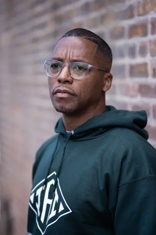 A man looking passed the camera in a dark green hoodie wearing glasses in front of a brick wall.
