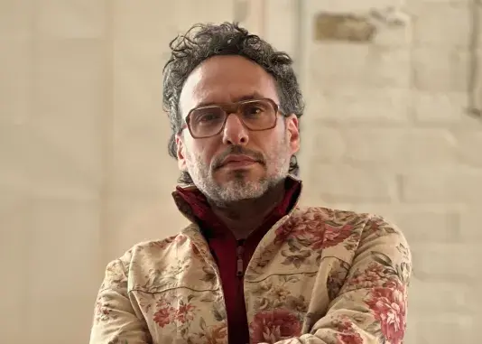 A portrait of Nida Sinnokrot, a man with a medium-light skin tone and short black and grey hair. He looks into the camera wearing eyeglasses and a cream jacket with pink floral accents.